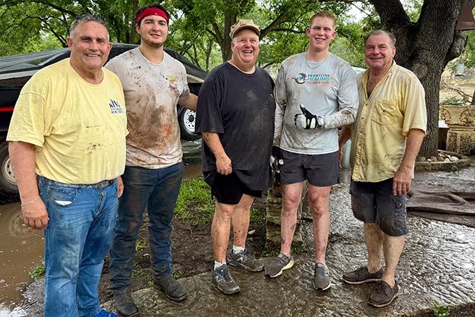 A group of five men, smiling proudly, stand outdoors on a wet pathway. They are covered in mud and appear to have been working, conveying a sense of camaraderie and accomplishment. Trees and a vehicle are visible in the background.