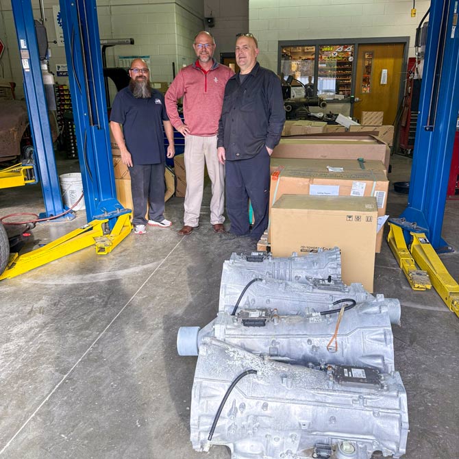 Three men stand smiling in a workshop, surrounded by car parts and boxes. A car lift and automotive equipment are visible, conveying a friendly, collaborative atmosphere.