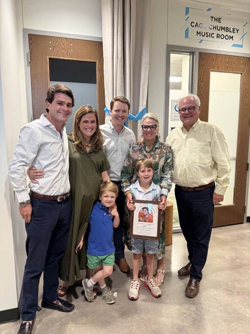 A family of seven, including two young children, poses happily in front of a music room. One child holds a framed photo, highlighting a commemorative moment.