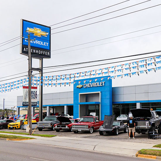 Atzenhoffer Chevrolet; A Chevrolet dealership with classic cars displayed in front. A blue sign with the Chevrolet logo is visible. Colorful flags hang above, creating a festive atmosphere.