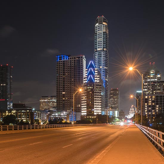 President’s Message: The Truth of Our Impact; Nighttime cityscape showing a well-lit road leading to illuminated skyscrapers under a dark sky in Austin. Bright blue accents add a modern touch.